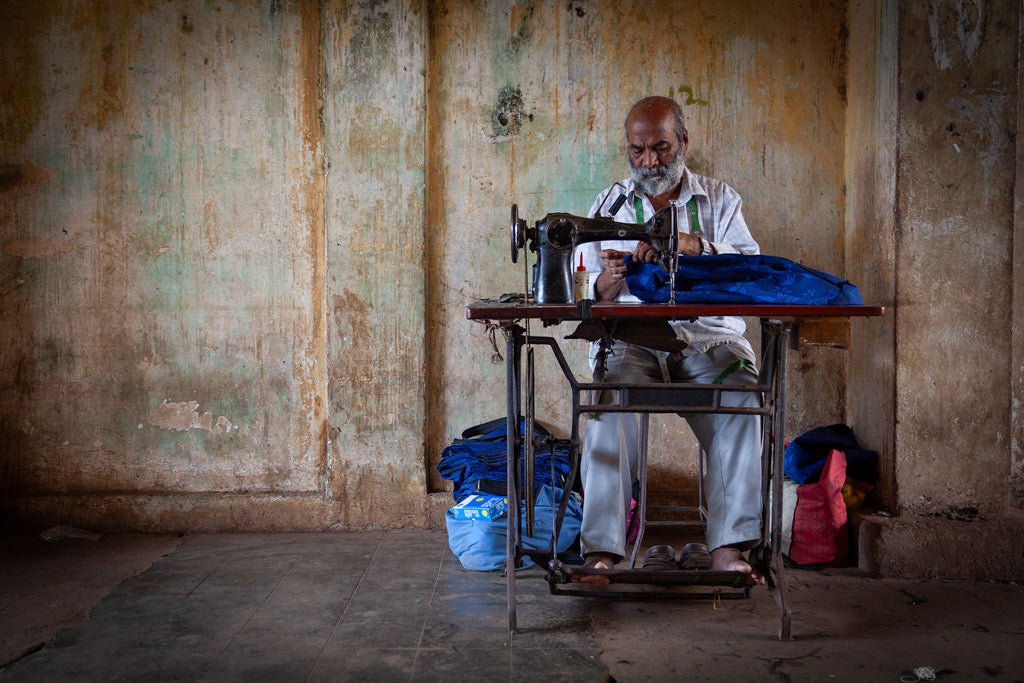 An elderly street seamster with a white beard sits at a vintage sewing machine, diligently working on blue fabric in a dimly lit room with worn, rustic walls and a tiled floor. Various bags are placed around him, adding to the authentic ambiance of Goan craftsmanship. This vivid scene captures the essence of *Street Seamster: Colour* by FN Prints.