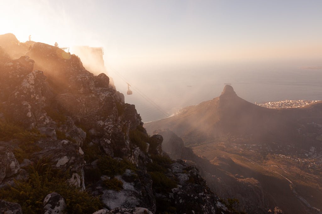 A mountain covered in rocky terrain and sparse vegetation is enveloped in a golden mist. In the distance, Table Mountain rises sharply from the landscape. The Table Mountain Cable Car by FN Prints can be seen traversing between the peaks, with the ocean and a coastal city visible below—a scene perfect for a photography print.