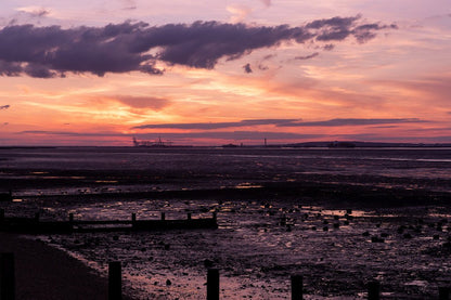 The "Thames Estuary Dusk" by FN Prints captures a serene sunset over the Thames Estuary tidal flat, where shades of purple, orange, and pink merge seamlessly in the sky. Silhouetted industrial structures and a shoreline appear on the distant horizon. The darkened, muddy flats in the foreground are dotted with scattered debris, resembling breathtaking coastal sunset artwork.