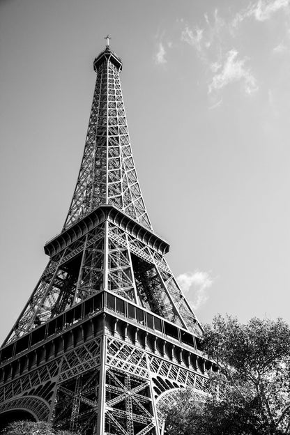 The Eiffel Tower: Part I by FN Prints beautifully captures the architectural splendor of the iconic iron lattice structure in a black and white photograph. Viewed from below against a partly cloudy sky, the image showcases its majesty with subtle hints of tree foliage at the base.