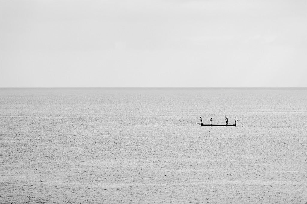 Minimalist fine art photography of a small fishing boat with three people on a calm, misty blue sea in Vietnam with no visible horizon.