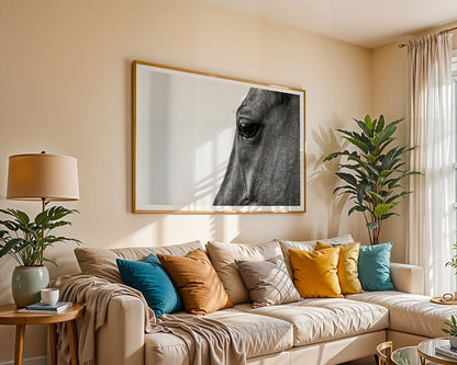 A cozy living room includes a beige sofa adorned with multi-colored cushions, accompanied by a wooden side table topped with a lamp and surrounded by potted plants. A large framed print titled "The Horse" by FN Prints is prominently displayed on the wall, as sunlight streams through sheer curtains.