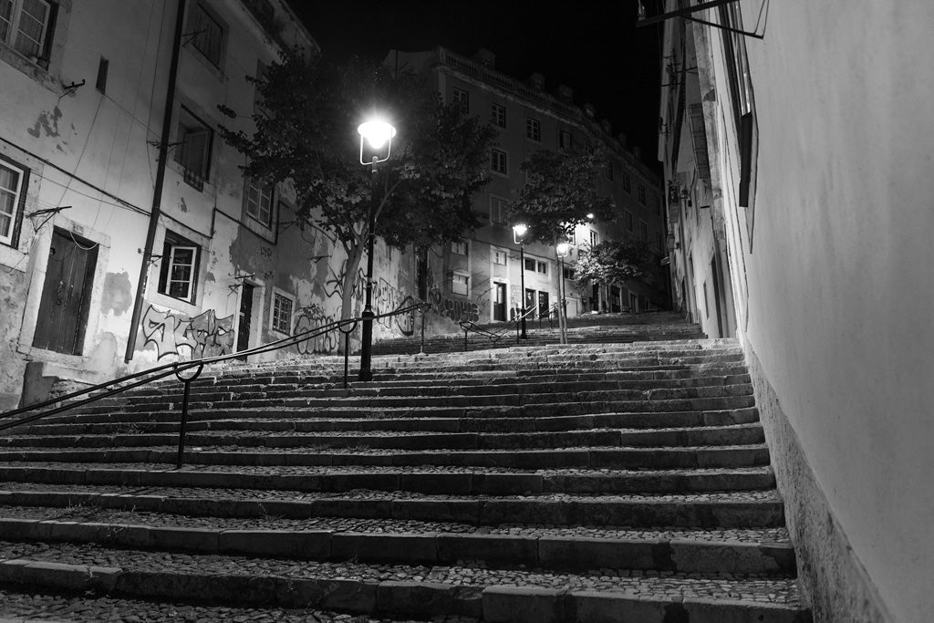 Product Description: "The Staircase" by FN Prints captures a steep, cobblestone staircase in a narrow alleyway at night, illuminated by street lamps. The old buildings lining the street bear graffiti on their walls, and a metal handrail runs up the middle of the steps. This black and white photograph also features trees visible at the top of the stairs.