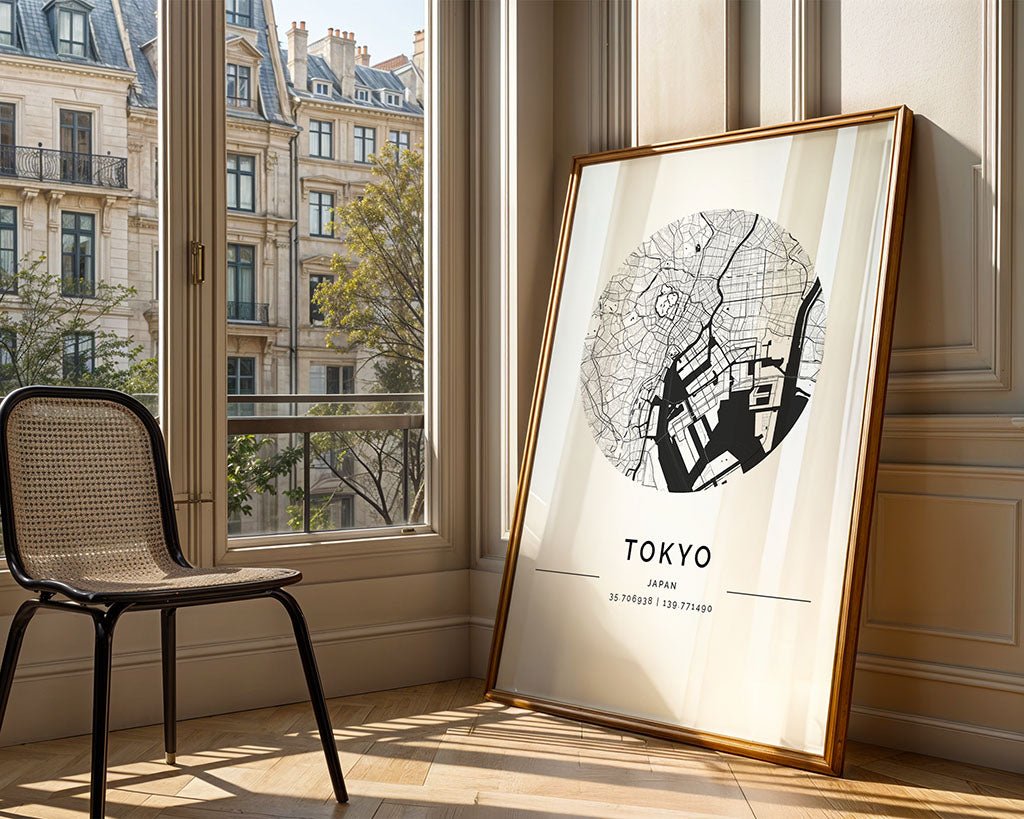 A framed FN Prints Tokyo City Map in premium black and white print leans against a wall by a window with European buildings. To its left, sunlight filters through a woven chair, casting gentle shadows, creating an elegant artful ambiance.