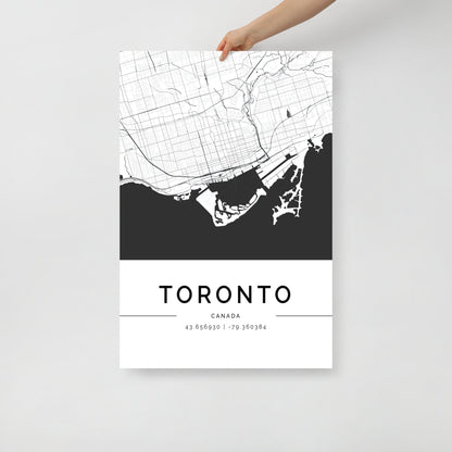 Someone is holding a Toronto Map poster by FN Prints, designed as a black and white wall art print. The map intricately showcases streets and waterfront details, with the coordinates "43.653900 | -79.360384" elegantly displayed below.