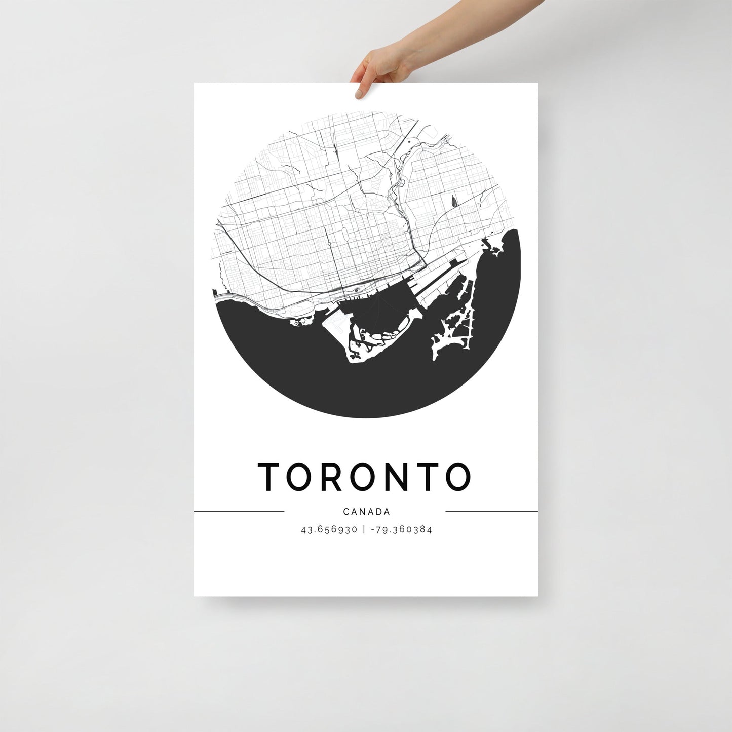 A person holds a stylish Toronto Map by FN Prints, showcasing a sleek black and white design of the city's layout. The poster includes geographic coordinates 43.653908, -79.384384 printed below and features a circular design ideal for wall art, capturing the essence of this vibrant Canadian city.