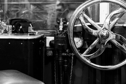 A black and white close-up image of the Tram Cab by FN Prints, showcasing industrial machinery with a large, shiny metal wheel centered by a bolt, multiple control levers, and a pressure gauge surrounded by various pipes and components. This captures an old-fashioned mechanical setup with vintage charm.