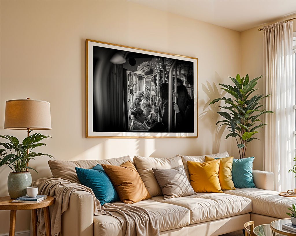 In a cozy living room, a light beige couch with colorful cushions is complemented by a floor lamp on the left. Hanging on the wall above is "Tram Tales," a large black-and-white framed photograph by FN Prints that captures the urban sophistication of San Francisco street life, featuring people seated in what appears to be a subway train.