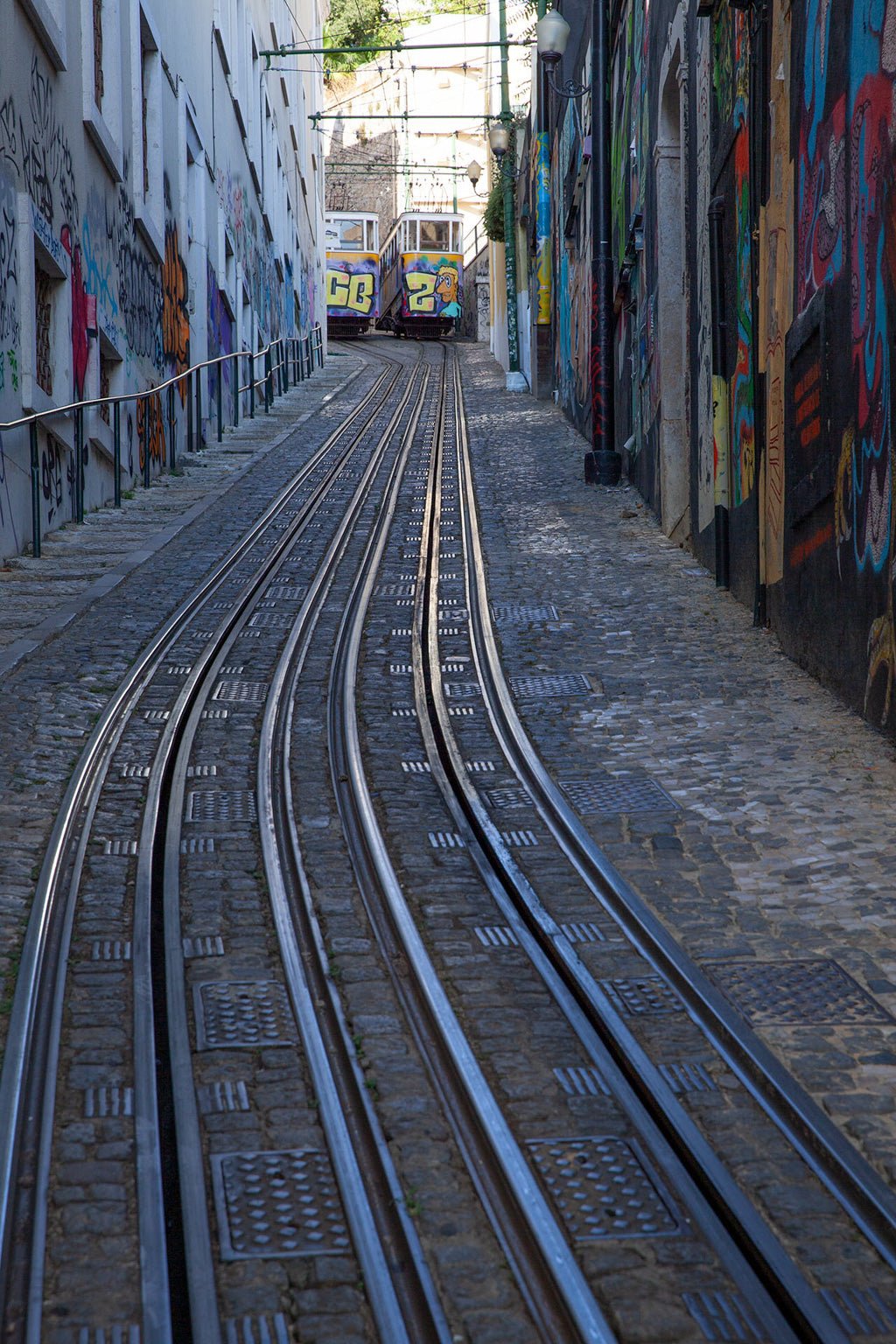 A steep street in the city showcases FN Prints' "Tram Tracks Colour" with two sets of funicular railway tracks, flanked by colorful graffiti on both sides. Two tram cars, painted in vibrant hues, are seen ascending the hill. The area is paved with cobblestones, enhancing the urban landscape art charm.