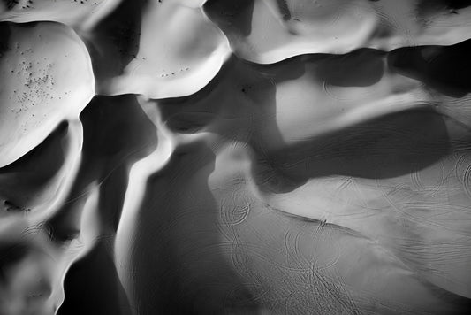 FN Prints' "Dune Part II Black & White" offers a captivating aerial photograph of a mesmerizing desert landscape. The sand dunes create smooth, wavy patterns with varying shades and shadows. Tire marks are visible on the sand, adding texture to the otherwise untouched surfaces.