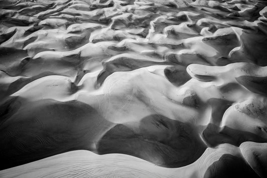 Dune Part IV Black & White by FN Prints captures the expansive sand dunes of the UAE desert in a stunning aerial black and white photograph. The intricate sand formations create mesmerizing patterns with smooth, wavy ridges, while faint tire tracks add texture to the vast, serene landscape.