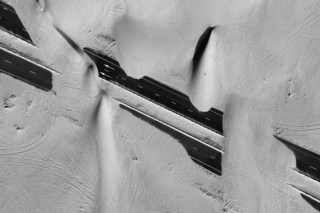 Aerial view of a snow-covered landscape with dark, straight lines of a road cutting through the white ground. Several patches of exposed road contrast sharply against the surrounding snow, forming abstract shapes and shadows reminiscent of FN Prints' stunning artwork, Desert Roads III.