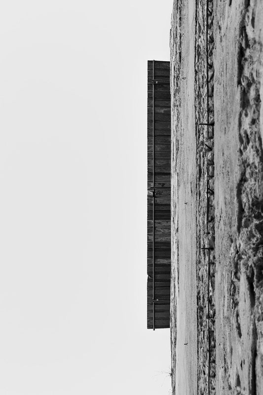 A black and white photograph titled "Up The Wall" by FN Prints showcases a dramatically tilted perspective of a stone building and an overhanging wooden structure, possibly a balcony or roof. The rustic charm is emphasized by the extreme angle, creating an almost abstract composition with stark contrast between the architectural details and the sky.