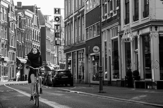 The FN Prints' Urban Cyclist artwork captures urban life in a black and white street photograph, showcasing a person riding a bicycle down a city street. Sporting a scarf and striped leggings, they skillfully weave through parked cars and street signs. Tall buildings with an Amsterdam-like charm line both sides of the road, all under the watchful presence of a vertical "HOTEL" sign.