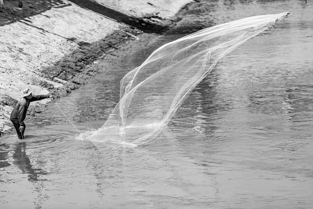 In a captivating black and white photograph titled "Urban Fisherman" by FN Prints, a Cambodian fisherman wearing a hat stands in shallow water near a concrete embankment in urban Cambodia. He is casting a large fishing net into the river, with the net mid-air creating an elegant arc above the water.