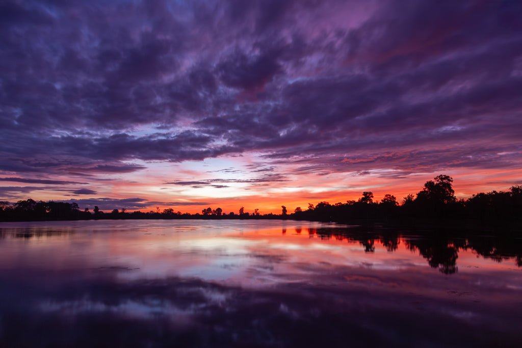 A serene lake reflecting a vibrant sunset with dramatic shades of purple, pink, orange, and red. The sky is filled with fluffy clouds, and silhouettes of trees and distant shoreline are visible against the colorful horizon—a perfect scene for "Vibrant Dawn," a photography print by FN Prints capturing natural serenity akin to a Cambodian sunrise.