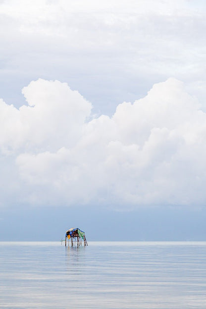 Minimalist fine art photography of a small stilt house floating on a calm sea under giant white clouds in Vietnam.