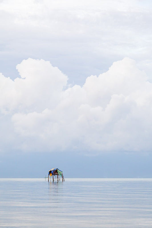 Minimalist fine art photography of a small stilt house floating on a calm sea under giant white clouds in Vietnam.