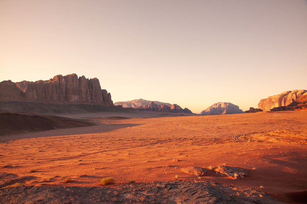 A vast desert landscape, bathed in warm light, stretches towards towering rock formations on the horizon. The terrain is sandy with sparse vegetation. The clear sky contrasts against the reddish-brown hues of the desert floor and distant mountains, evoking a Wadi Rum, Jordan scene by FN Prints.