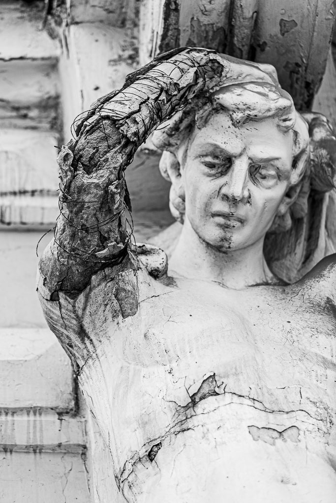 A black and white close-up photo titled "Weathered Strength" by FN Prints features a weathered stone sculpture in Ukraine, depicting a male figure with a partially lifted arm. The sculpture exhibits visible signs of decay and damage, including cracks, stains, and surface deterioration.