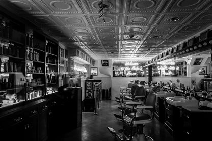 A black and white photograph, titled "Welcome To The Baba's House" by FN Prints, captures a vintage barber shop with retro flair. The space features a row of classic barber chairs, illuminated mirrors, and shelves stocked with grooming products. Neon signs reading "Welcome to Baba's House" add a nostalgic glow.