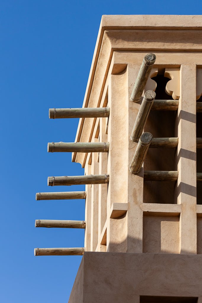 A close-up of FN Prints' Windcatcher extending from a beige clay building against a clear blue sky. Multiple wooden poles protrude horizontally from the tower's side, embodying Arabian architecture and designed to direct wind downwards for natural cooling—an early form of sustainable architecture.